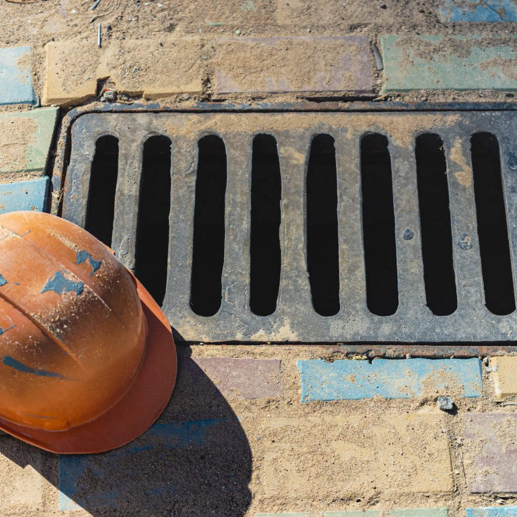 A cast-iron stormwater drainage hatch on the sidewalk with a construction helmet lying on it. Collection and drainage of rainwater from the street